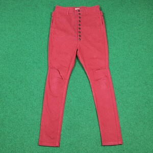 May include: Red denim pants with a button-up fly. The pants have a high waist and a slim fit. The knees have a distressed look. The pants are made of a sturdy denim material.