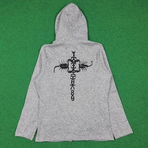 May include: A light gray hooded sweatshirt with a black graphic design on the back. The design features a cross-like shape with scorpion-like figures and stylized symbols. The sweatshirt has a full zipper and a hood.