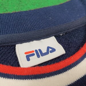 Puede incluir: Primer plano de una sudadera azul marino con el nombre de la marca "FILA" en blanco y rojo en una etiqueta. El cuello presenta rayas rojas y blancas. La tela tiene un aspecto texturizado.