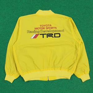 May include: Bright yellow bomber jacket with a ribbed collar and cuffs. The back displays the text "TOYOTA MOTOR SPORTS Racing Development TRD" in black and red. The jacket has a full-length zip and is made of a lightweight material.