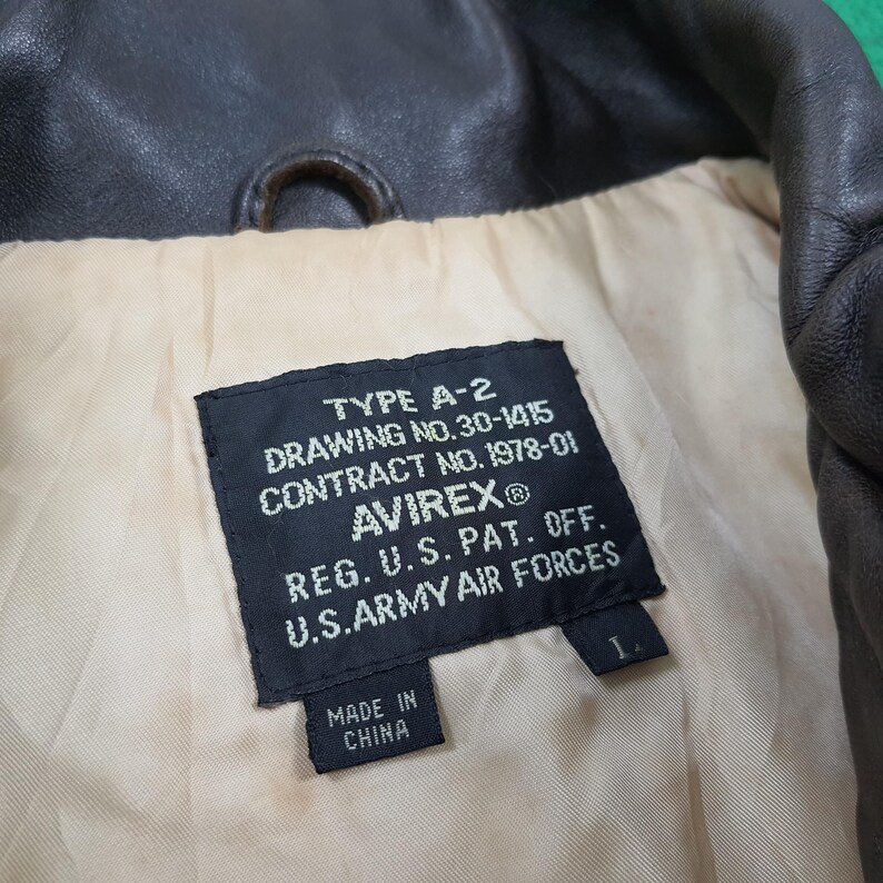 Puede incluir: El interior de una chaqueta de cuero marr&oacute;n oscuro con una etiqueta rectangular negra con texto blanco que dice TYPE A-2, DRAWING NO. 30-1415, CONTRACT NO. 1978-01, AVIREX, REG. U.S. PAT. OFF., U.S. ARMY AIR FORCES, MADE IN CHINA y una etiqueta L.