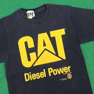 May include: A black short-sleeved t-shirt featuring a large yellow CAT logo on the chest. Below the logo, the text reads Diesel Power, with additional small text including Caterpillar Inc. 1993 and a circular CAT logo. A white CAT brand tag is at the neck.