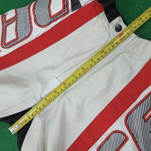 May include: Close-up of a section of a white and red racing jacket. The jacket has black and white striped accents and a red stripe. A yellow tape measure is laid across the jacket, showing measurements in inches.