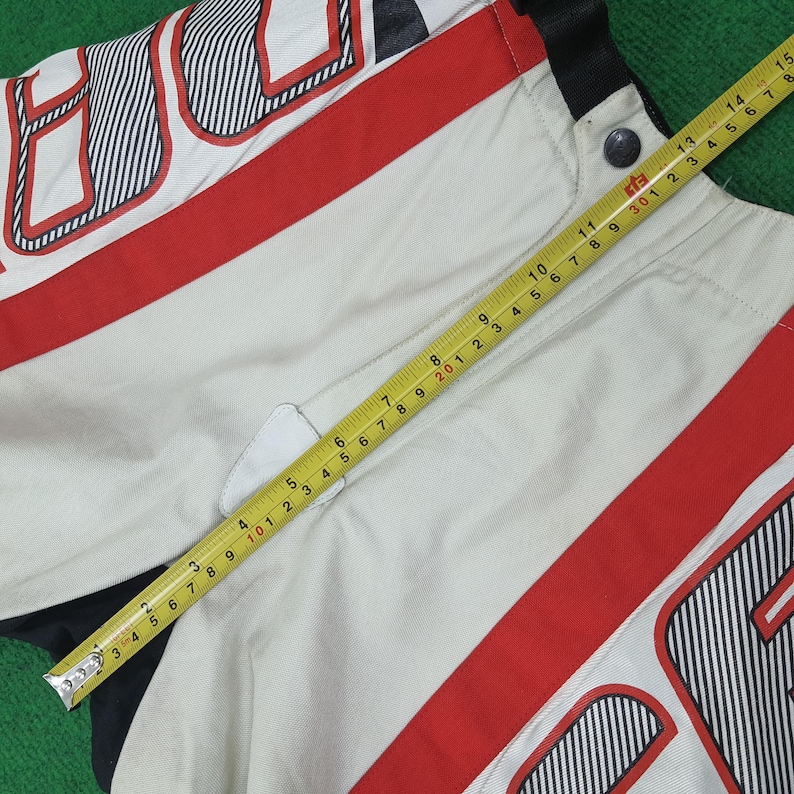 May include: Close-up of a section of a white and red racing jacket. The jacket has black and white striped accents and a red stripe. A yellow tape measure is laid across the jacket, showing measurements in inches.