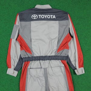Puede incluir: Mono de carreras Toyota gris y rojo. El mono presenta el logotipo de Toyota en la espalda, con detalles en gris, rojo y negro. Tiene cierre de cremallera y múltiples bolsillos. Diseñado para carreras o trabajo automotriz.