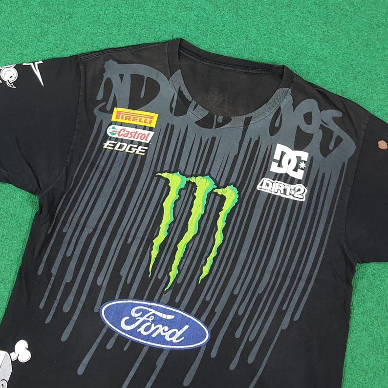 May include: A black short-sleeve t-shirt featuring a large green Monster Energy logo, a blue Ford oval logo, and various sponsor graphics including Pirelli, Castrol Edge, and DC Dirt 2. The shirt has a black drip pattern graphic across the chest.