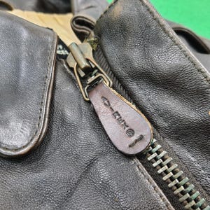Puede incluir: Una chaqueta de cuero marr&oacute;n oscuro con cierre de cremallera met&aacute;lica y un tirador de cuero a juego. El tirador tiene grabado el texto 'EXCEL' y un peque&ntilde;o s&iacute;mbolo. La chaqueta presenta un acabado de grano texturizado y forro interior beige.