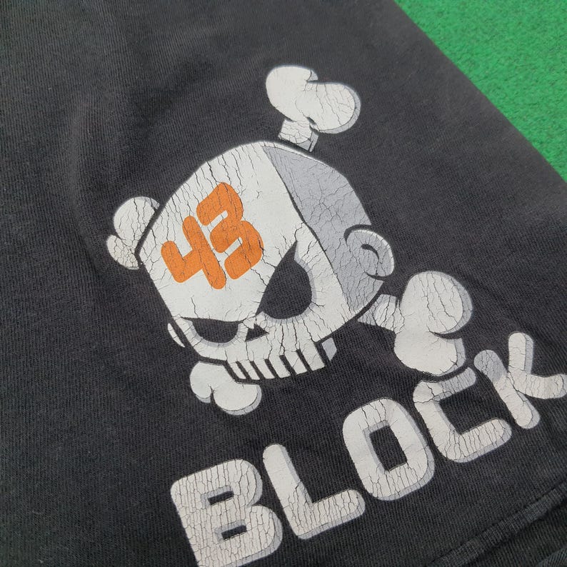 May include: A black t-shirt featuring a graphic of a stylized skull and crossbones design. The skull is light gray with a prominent orange number 43 printed on the forehead. Below the graphic, the word BLOCK is printed in large white letters.