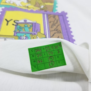May include: White fabric with a green tag and colorful stamp-like designs. The stamps feature illustrations of bears, a theater scene, and other graphics. The tag has handwritten numbers and letters.