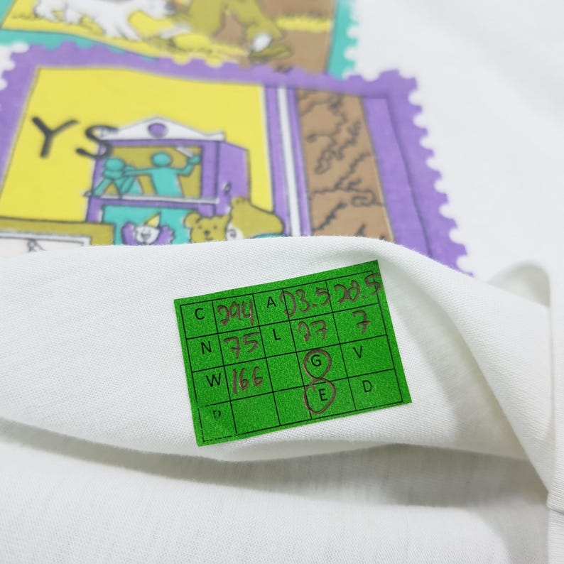 May include: White fabric with a green tag and colorful stamp-like designs. The stamps feature illustrations of bears, a theater scene, and other graphics. The tag has handwritten numbers and letters.