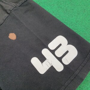 May include: A black t-shirt featuring a large white number 43 printed on the lower portion. The white graphic shows visible signs of cracking in the ink. Additionally, there is a small, circular brown stain located on the fabric near the number.