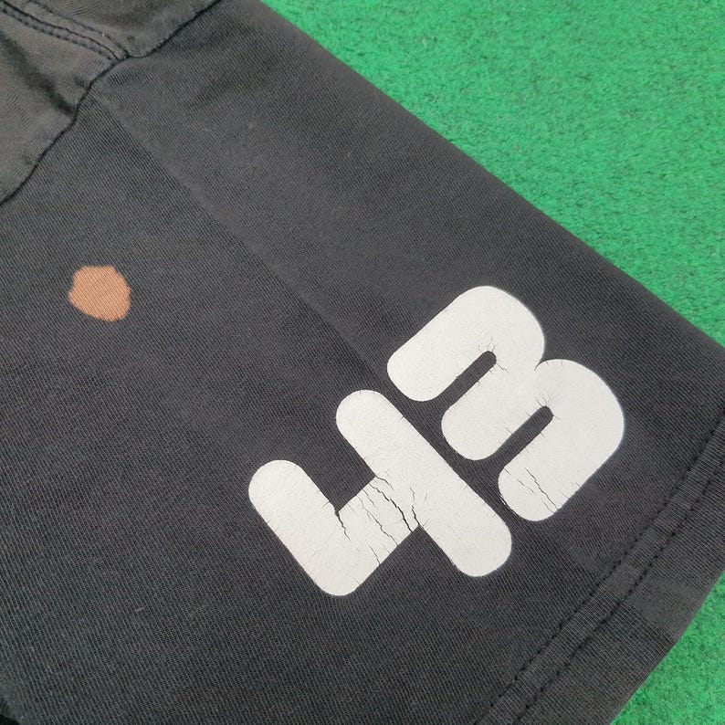 May include: A black t-shirt featuring a large white number 43 printed on the lower portion. The white graphic shows visible signs of cracking in the ink. Additionally, there is a small, circular brown stain located on the fabric near the number.