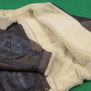 Puede incluir: Chaqueta de cuero marr&oacute;n con forro de borrego color crema. La chaqueta presenta cierre de cremallera y un estilo aviador cl&aacute;sico. Las mangas tienen parches reforzados, y el dise&ntilde;o sugiere calidez y durabilidad. La chaqueta est&aacute; sobre una superficie verde.