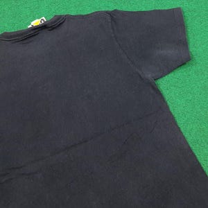 May include: A solid black short-sleeved t-shirt featuring a crew neckline and a small white and yellow brand tag attached to the inside of the collar. The garment is plain with no additional graphics or patterns visible on the fabric surface.