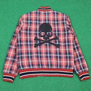 Könnte beinhalten: Rote, weiße und blaue karierte Bomberjacke mit schwarzem Totenkopf- und gekreuzten Knochen-Motiv auf dem Rücken. Die Jacke hat einen gerippten Kragen, Bündchen und Saum mit rot-weiß-blauen Streifen. Die Jacke wird auf grünem Hintergrund gezeigt.