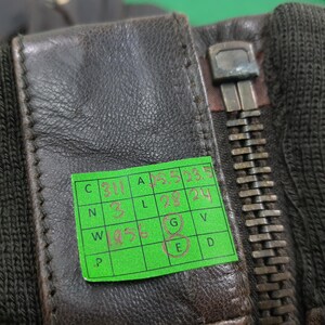 Puede incluir: Una chaqueta de cuero marr&oacute;n oscuro con cremallera met&aacute;lica y pu&ntilde;os de punto acanalado negro. Una pegatina de cuadr&iacute;cula verde est&aacute; adherida al cuero, mostrando medidas y c&oacute;digos escritos a mano en tinta roja, incluyendo 23.5, 28, 24 y 1956.