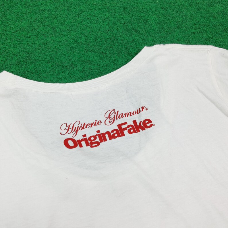Puede incluir: Una camiseta blanca con texto rojo impreso en la parte superior de la espalda. El texto dice Hysteric Glamour en una fuente cursiva sobre la palabra OriginaFake en una fuente negrita sin remates. La prenda tiene un cuello redondo sencillo.