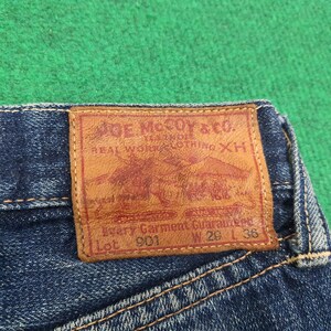 K&ouml;nnte beinhalten: Nahaufnahme eines blauen Jeans-Etiketts. Das braune Lederetikett tr&auml;gt die Aufschrift "JOE McCOY & CO." mit einer Grafik eines Pferdewagens. Das Etikett enth&auml;lt auch "W 29 L 36" und "Every Garment Guaranteed".