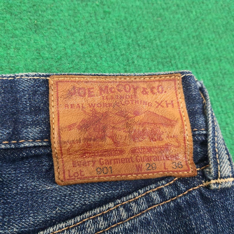 K&ouml;nnte beinhalten: Nahaufnahme eines blauen Jeans-Etiketts. Das braune Lederetikett tr&auml;gt die Aufschrift "JOE McCOY & CO." mit einer Grafik eines Pferdewagens. Das Etikett enth&auml;lt auch "W 29 L 36" und "Every Garment Guaranteed".