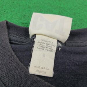 May include: A dark gray t-shirt featuring a white interior neck label with the letters CML printed in large font. The tag includes care instructions, 100% cotton material, Made in U.S.A. text, and a size medium label on the fabric.