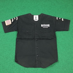 May include: Black short-sleeved button-up shirt with two chest pockets. Features patches with text and a US flag on the left sleeve. The shirt has the text "BAPE" on the chest and a small logo.