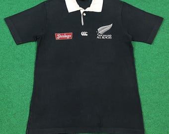 Puede incluir: Camiseta de rugby negra con cuello blanco y mangas cortas. Presenta el logo "Steinlager" en el pecho izquierdo, el logo Canterbury y el logo "NEW ZEALAND ALL BLACKS" con un helecho en el pecho derecho.