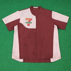 Puede incluir: Una camisa de uniforme de manga corta burdeos y blanca con el logo "7-Eleven" en el pecho. La camisa tiene cuello mandarín, dos bolsillos delanteros y un panel blanco contrastante en el lado izquierdo.