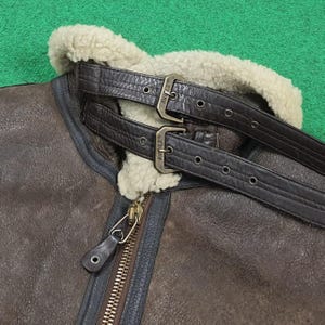 Puede incluir: Primer plano de una chaqueta de cuero marr&oacute;n con cuello de borrego crema. La chaqueta presenta dos correas de cuero marr&oacute;n con hebillas met&aacute;licas y una cremallera de lat&oacute;n. Chaqueta de estilo cl&aacute;sico.