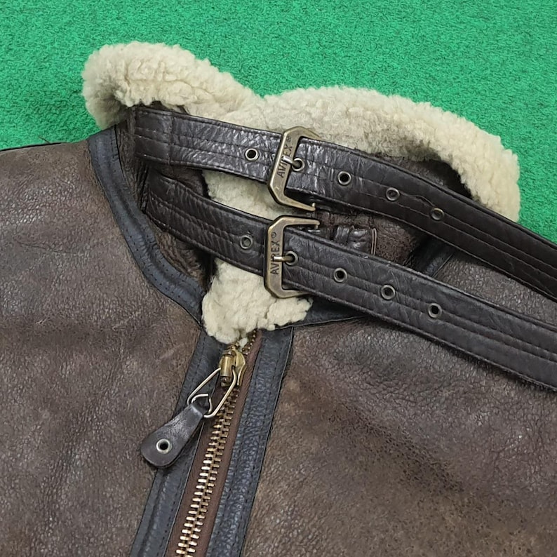 Puede incluir: Primer plano de una chaqueta de cuero marr&oacute;n con cuello de borrego crema. La chaqueta presenta dos correas de cuero marr&oacute;n con hebillas met&aacute;licas y una cremallera de lat&oacute;n. Chaqueta de estilo cl&aacute;sico.