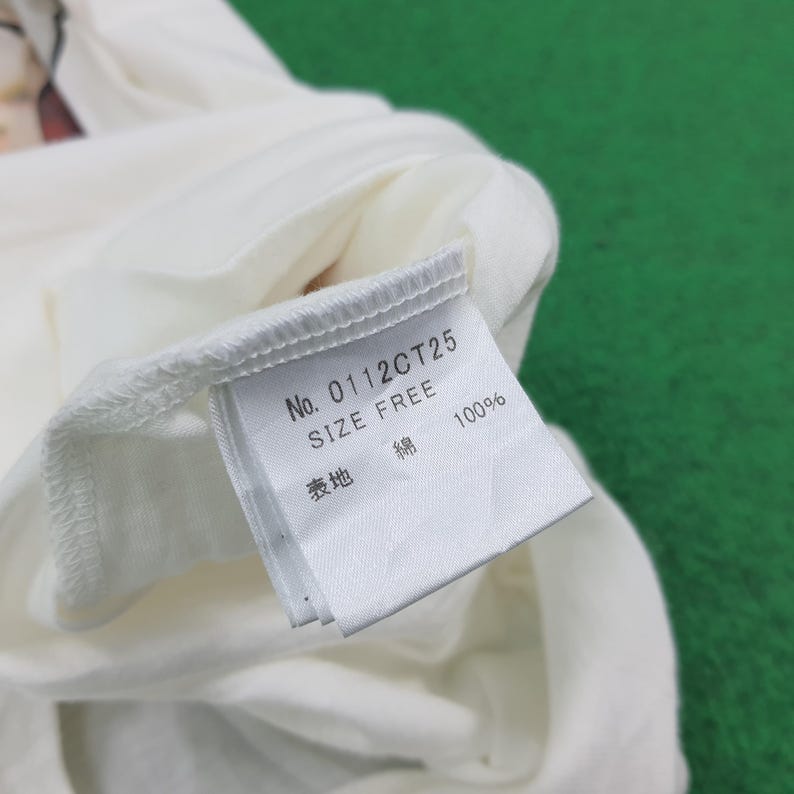 Puede incluir: Una prenda blanca con una etiqueta de cuidado cosida. La etiqueta muestra el texto No. 0112CT25, SIZE FREE y 100% en tinta negra. Tambi&eacute;n incluye caracteres japoneses que indican la composici&oacute;n de algod&oacute;n y el tejido exterior de la prenda.