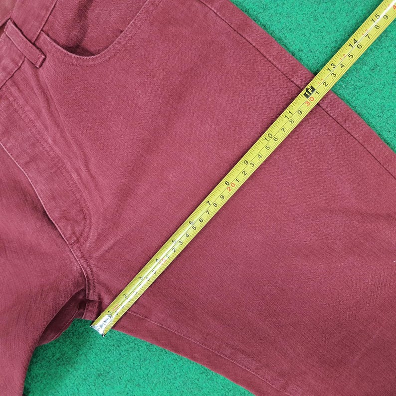 May include: Close-up of burgundy-colored denim pants with a yellow measuring tape. The tape shows measurements in inches. The pants are on a green surface. The pants have a classic five-pocket design.