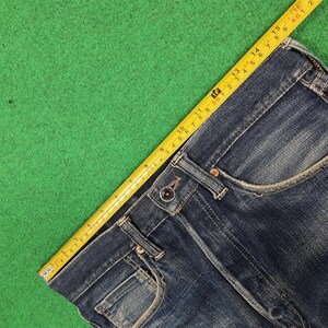 K&ouml;nnte beinhalten: Nahaufnahme einer blauen Jeans mit Knopfverschluss und Taschen. Ein gelbes Ma&szlig;band liegt &uuml;ber der Jeans und zeigt die Ma&szlig;e in Zentimetern an. Die Jeans liegt auf einer gr&uuml;nen Oberfl&auml;che.