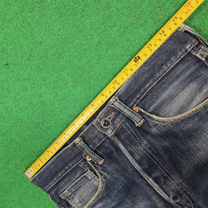 K&ouml;nnte beinhalten: Nahaufnahme einer blauen Jeans mit Knopfverschluss und Taschen. Ein gelbes Ma&szlig;band liegt &uuml;ber der Jeans und zeigt die Ma&szlig;e in Zentimetern an. Die Jeans liegt auf einer gr&uuml;nen Oberfl&auml;che.