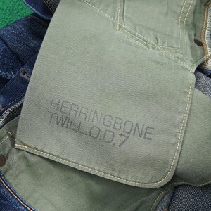 K&ouml;nnte beinhalten: Nahaufnahme einer Jeans mit gr&uuml;nem Taschenfutter. Das Taschenfutter tr&auml;gt den Text "HERRINGBONE TWILL O.D.7" in Dunkelgrau. Die Jeans ist blau mit gelben N&auml;hten.