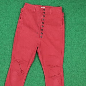 May include: Red pants with a high waist and a button-up fly. The pants have a classic design with a straight leg and pockets. The fabric appears to be a sturdy cotton or blend. The pants are displayed on a green surface.