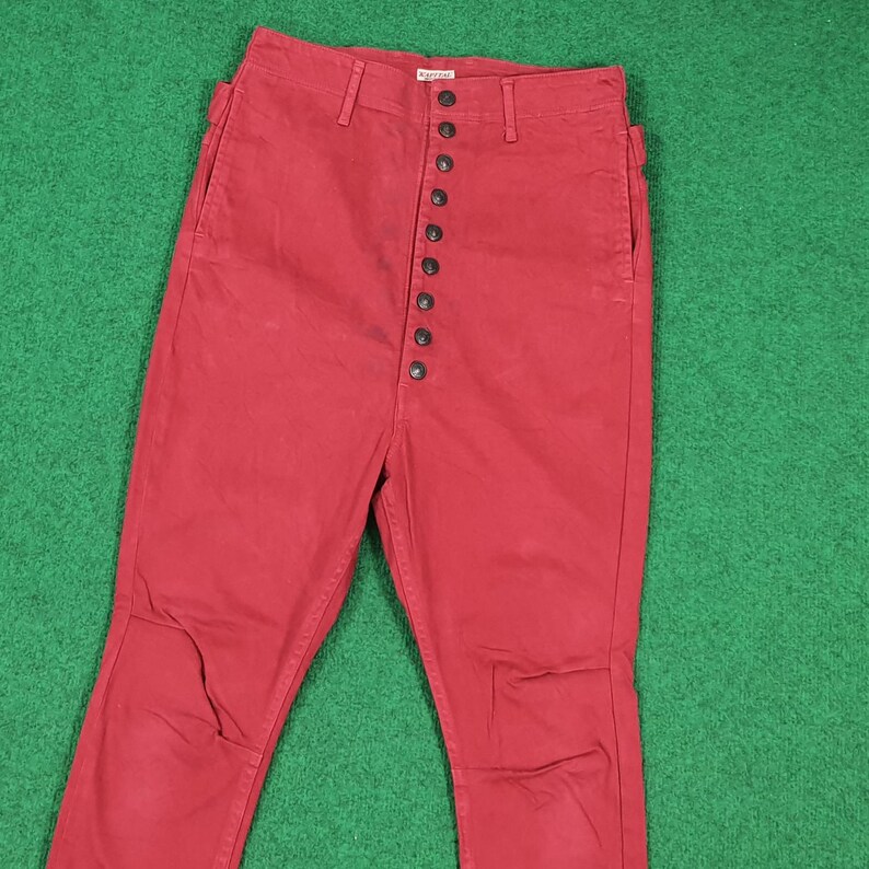 May include: Red pants with a high waist and a button-up fly. The pants have a classic design with a straight leg and pockets. The fabric appears to be a sturdy cotton or blend. The pants are displayed on a green surface.
