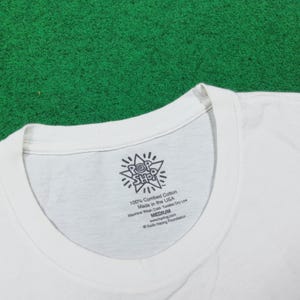 Puede incluir: Camiseta blanca con cuello redondo. La etiqueta muestra el texto "POP SHOP" y "100% Combed Cotton Made in the USA". La camiseta es de talla mediana. El fondo es verde.