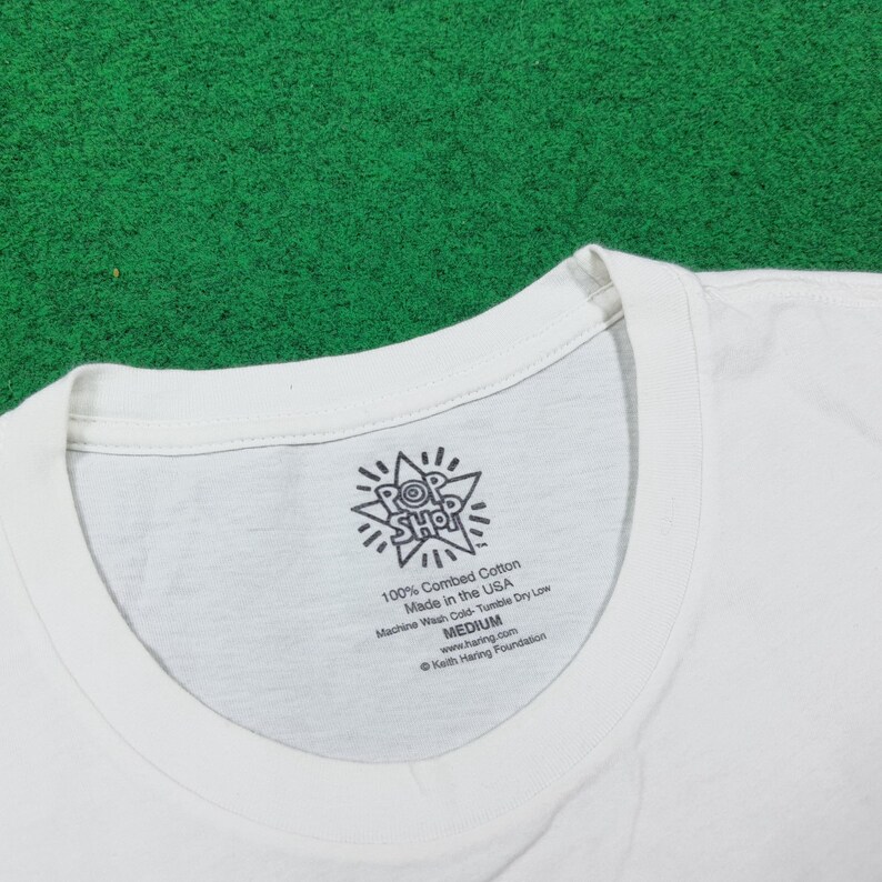 Puede incluir: Camiseta blanca con cuello redondo. La etiqueta muestra el texto "POP SHOP" y "100% Combed Cotton Made in the USA". La camiseta es de talla mediana. El fondo es verde.