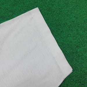 May include: A corner of a white fabric item, possibly a shirt or cloth, is displayed against a green background. The fabric appears to be slightly textured, with visible seams along the edges. The overall presentation is simple and clean.