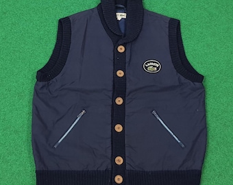 May include: Navy blue vest with a shawl collar and wooden buttons. Features a small embroidered Lacoste logo on the chest. Includes two zippered pockets and a ribbed knit trim.