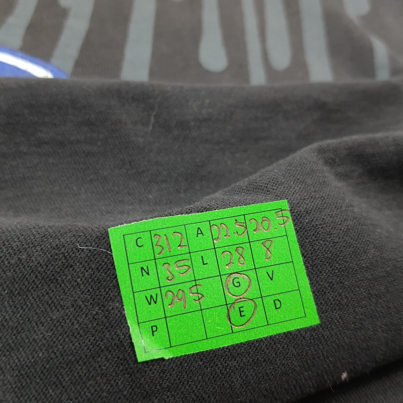 May include: A black fabric garment featuring a dark grey vertical stripe pattern and a bright green rectangular tag with a grid of handwritten measurements including 22.5, 20.5, 35, 28, 8, and 29.5, with the letters G and E circled in red ink.