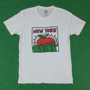 Puede incluir: Camiseta blanca con un dise&ntilde;o gr&aacute;fico. El dise&ntilde;o presenta el texto "NEW YORK" sobre una manzana roja con hojas verdes y l&iacute;neas radiantes. Figuras verdes est&aacute;n debajo de la manzana. La camiseta est&aacute; sobre una superficie verde.