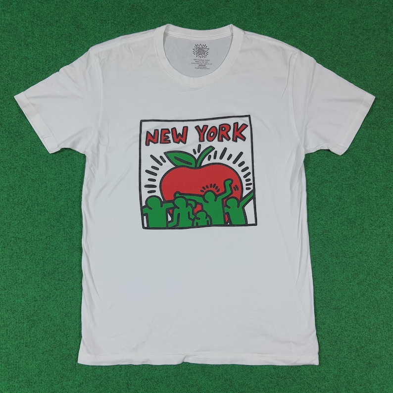 Puede incluir: Camiseta blanca con un dise&ntilde;o gr&aacute;fico. El dise&ntilde;o presenta el texto "NEW YORK" sobre una manzana roja con hojas verdes y l&iacute;neas radiantes. Figuras verdes est&aacute;n debajo de la manzana. La camiseta est&aacute; sobre una superficie verde.