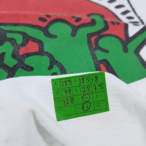 Puede incluir: Camiseta blanca con un dise&ntilde;o gr&aacute;fico verde y rojo. Una etiqueta verde con n&uacute;meros y letras manuscritos est&aacute; adjunta. El dise&ntilde;o es una figura estilizada con contornos negros.