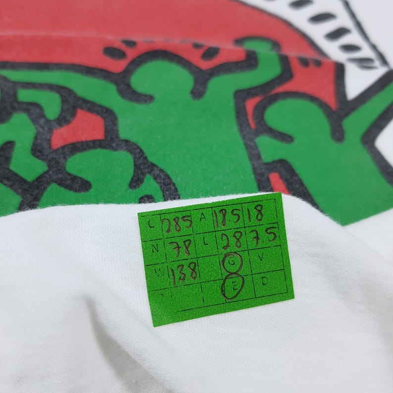 Puede incluir: Camiseta blanca con un dise&ntilde;o gr&aacute;fico verde y rojo. Una etiqueta verde con n&uacute;meros y letras manuscritos est&aacute; adjunta. El dise&ntilde;o es una figura estilizada con contornos negros.