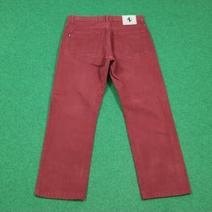 May include: A pair of burgundy-colored denim jeans. The jeans have a classic five-pocket design and a straight leg cut. A small white tag with a black horse logo is visible on the back pocket. The jeans are laid flat on a green surface.