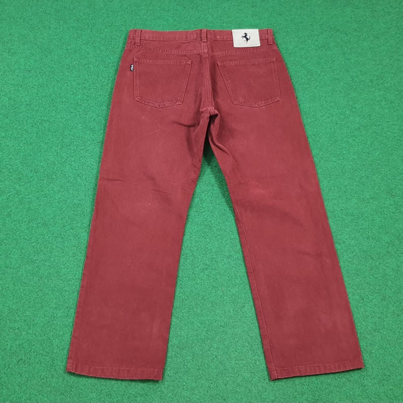 May include: A pair of burgundy-colored denim jeans. The jeans have a classic five-pocket design and a straight leg cut. A small white tag with a black horse logo is visible on the back pocket. The jeans are laid flat on a green surface.