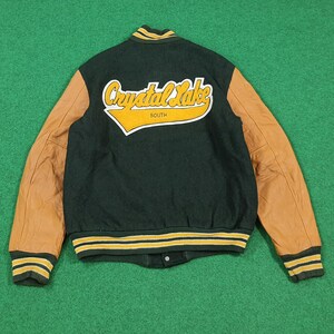 May include: A dark green and tan varsity jacket with the words "Crystal Lake South" in a yellow and white design on the back. The jacket has tan leather sleeves and green and yellow striped cuffs and collar.