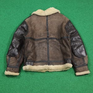 Puede incluir: Chaqueta de cuero marr&oacute;n con parches de cuero negro en los hombros y las mangas. La chaqueta tiene un cuello y un ribete de piel de oveja de color crema. La chaqueta est&aacute; sobre una superficie verde.