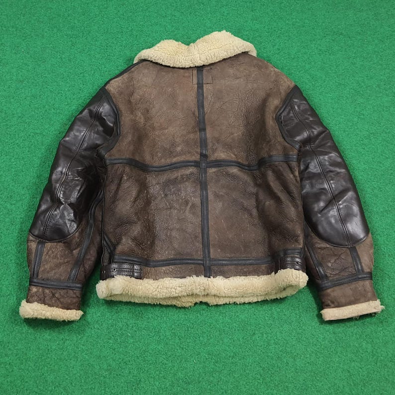 Puede incluir: Chaqueta de cuero marr&oacute;n con parches de cuero negro en los hombros y las mangas. La chaqueta tiene un cuello y un ribete de piel de oveja de color crema. La chaqueta est&aacute; sobre una superficie verde.